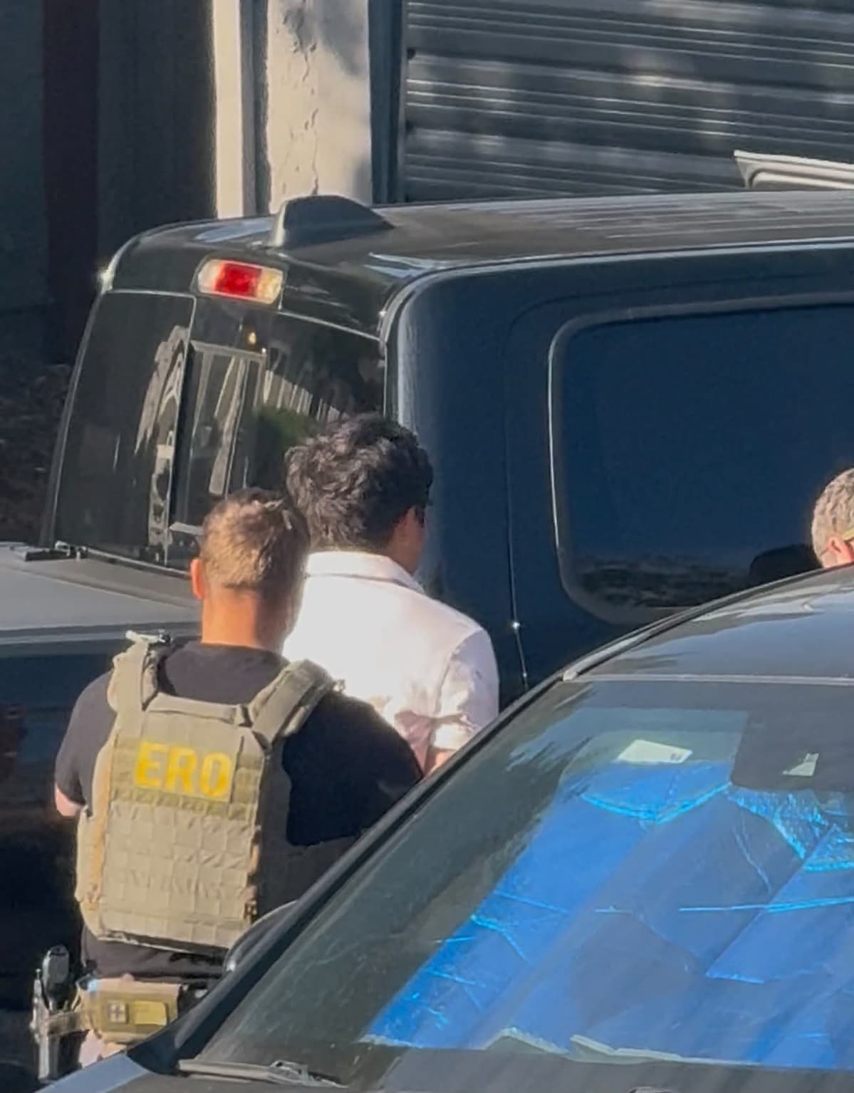 An agent whose vest reads "ERO," Enforcement and Removal Operations, the ICE division responsible for immigration arrests and deportations, walks a man in a white polo shirt across the lot.