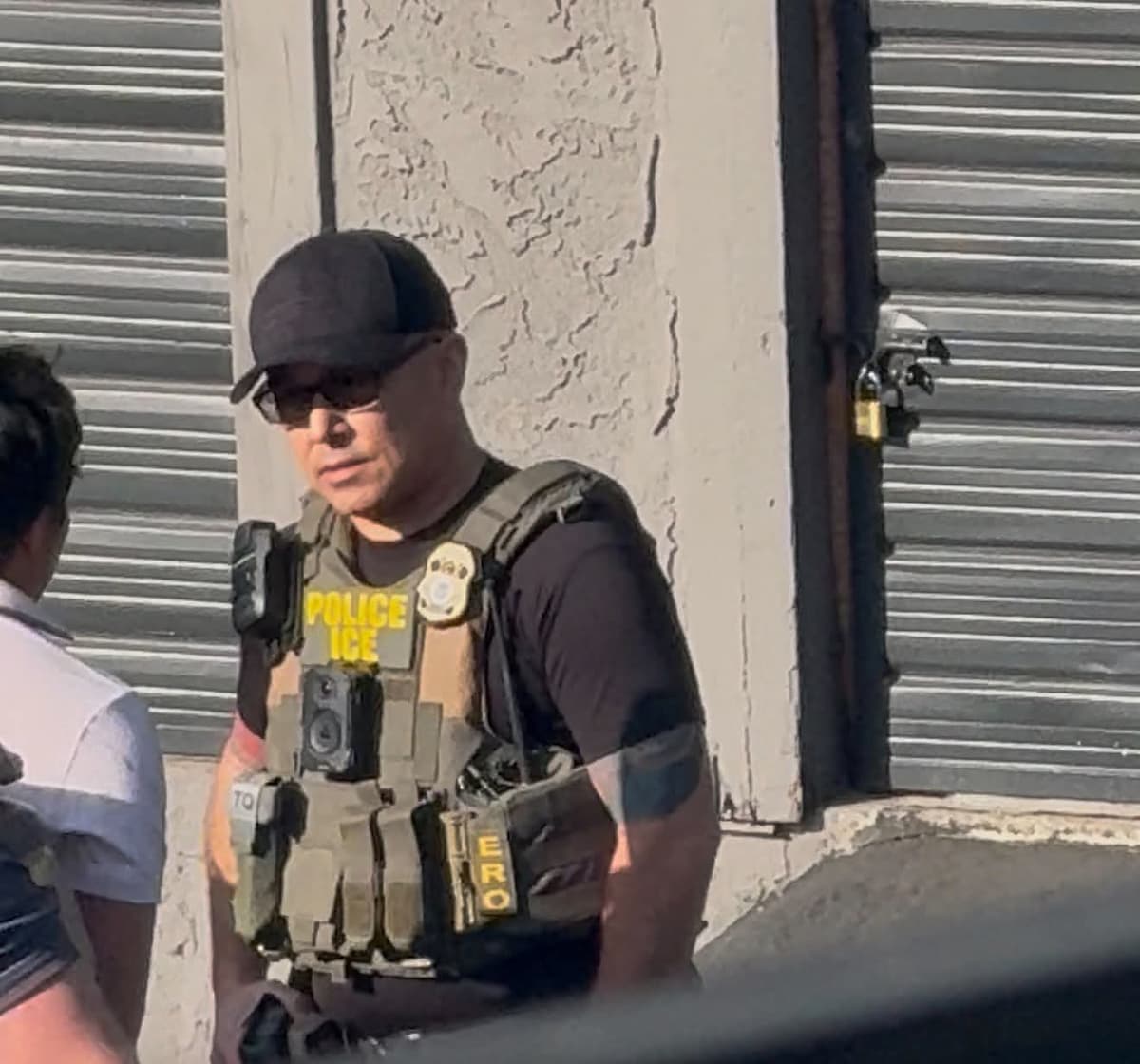An agent in a vest marked "POLICE ICE" wears a badge and two body-worn cameras at The Peaks Apartments on Churn Creek Road in Redding. No name tape is visible.